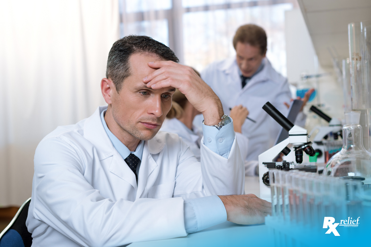 A stressed man in a white lab coat puts his hand on his forehead in frustation. He is sitting at a lab station with a microscope and test tubes filled with red liquid in them. His co-workers yells at someone in the background angrily.