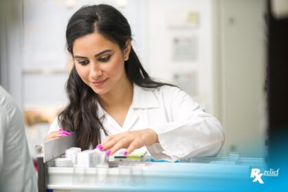 Female Middle Eastern pharmacist in white lab coat looks at boxes of medicine in an open drawer.